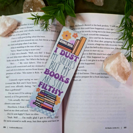 Book with a bookmark titled 'I Dust My Shelves But My Books Are Still Filthy' on an open book, surrounded by decorative elements.